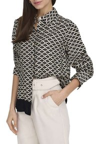 Blouse à motifs noir et blanc avec un col, à demi-boutonnée, présentant des manches longues, un ourlet noir uni, et une coupe décontractée, associée à un pantalon crème.
