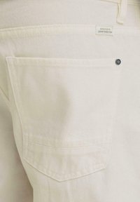 Κοντινή λήψη μιας πίσω τσέπης από εκρού τζιν με μια μικρή μαύρη τριχιά και μια ετικέτα που γράφει «Αφιερωμένο στην τελειότητα του τζιν».