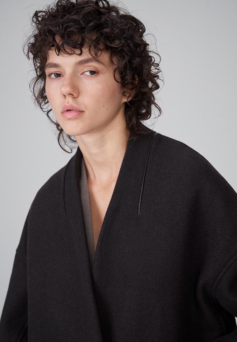 Giovane con i capelli ricci corti che indossa un cappotto scuro largo, che guarda leggermente di lato su uno sfondo chiaro e uniforme.