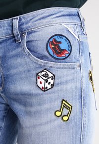 Jeans i denim med blå tvätt, prydda med färgglada broderade lappar inklusive röda högklackade skor, en tärning och musiknoter på framsidan.