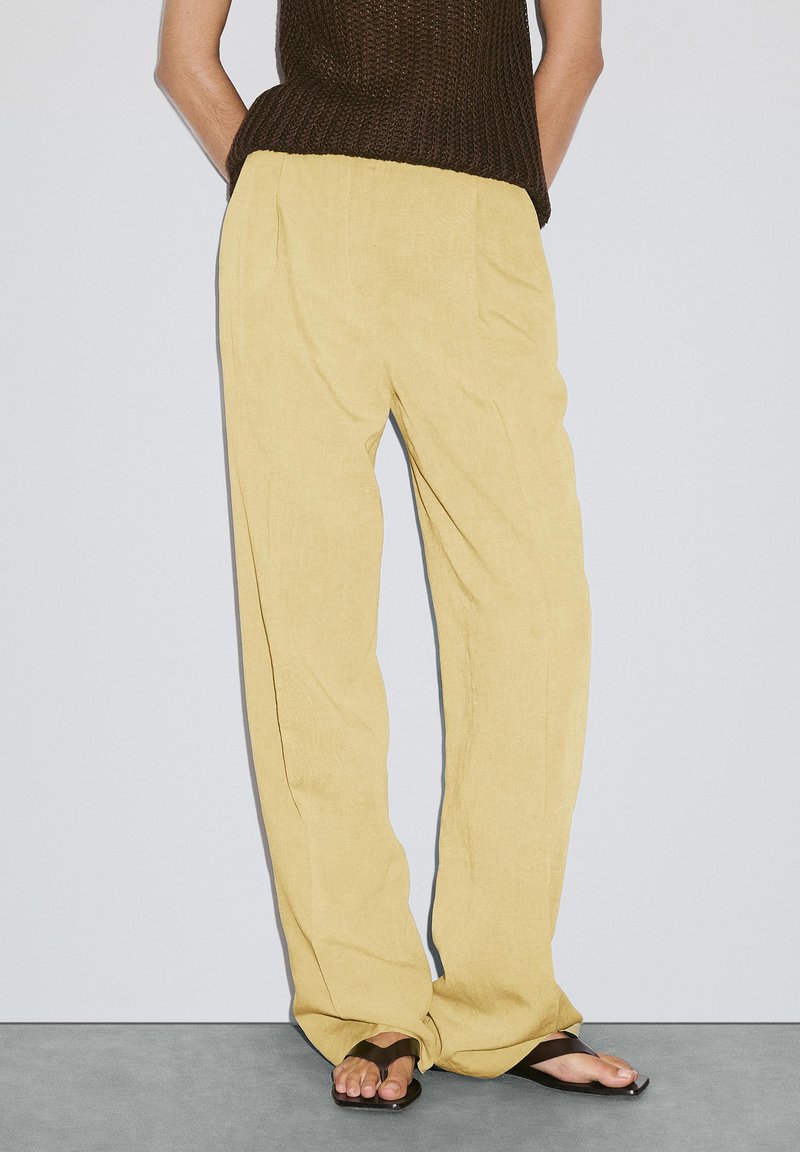 Lichtgele linnen broek met een ontspannen fit en voorplooien, gecombineerd met donkerbruine flip-flops op een grijze ondergrond.