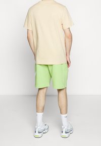 Jasnobeżowa koszulka z krótkim rękawem o luźnym kroju, zestawiona z neonowymi zielonymi szortami o gładkiej fakturze. Białe skarpetki sportowe i szare trampki.