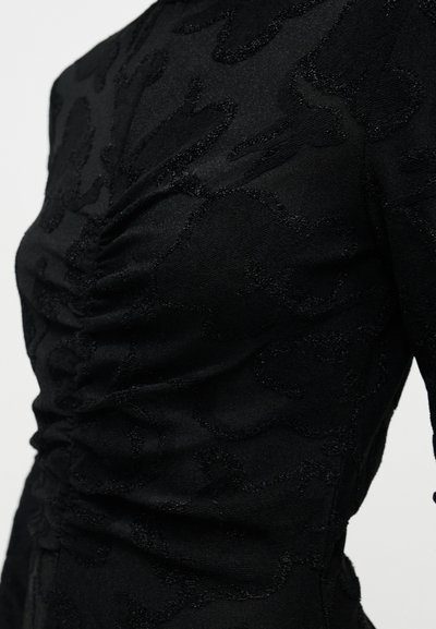 Fekete felső, texturált anyagból, dombornyomott mintával. A kialakítás oldalán ráncolt részletekkel és testhezálló sziluettel rendelkezik.