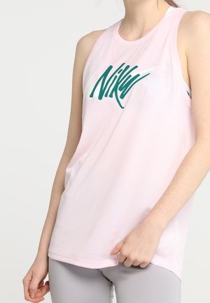Vrouw draagt een lichtroze mouwloze Nike top met groen logo, gecombineerd met lichtgrijze legging, hand rustend bij de heup.