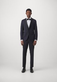 Dunkelblaues Smoking mit schimmerndem Schalkragen, weißem Hemd, schwarzer Fliege und schwarzen Lederschuhen. Maßgeschneiderter Slim-Fit, klassisches Design.