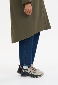 Person trägt olivgrünen Mantel, blaue Jeans, weiße Socken und beige-schwarze Wanderschuhe und steht vor weißem Hintergrund.