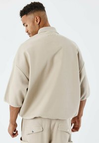 Camicia beige oversize in tessuto testurizzato, con logo sul colletto posteriore e spalle scese per un fit casual. Abbinata a pantaloni coordinati.