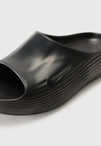 Melna slide čība, kas izgatavota no gludas plastmasas, ar izliekto augšdaļu un atvērtu purngalu. Iezīmēta ar rievojumu un melnu Nike logo.