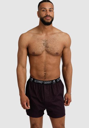 Shirtless man with short beard and earrings wearing dark boxer shorts with "LOUSY LIVIN" waistband, standing against plain light background.