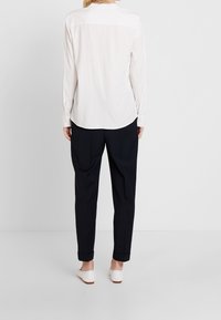 Blouse à manches longues beige clair avec un col rond, une texture lisse et un détail plissé dans le dos, associée à un pantalon noir fuselé.
