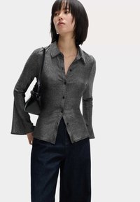 Skinnende sort skjorte med lange, flagrende ærmer, krave-design og diskret glimmer, parret med mørke denim bukser og en lille sort taske.