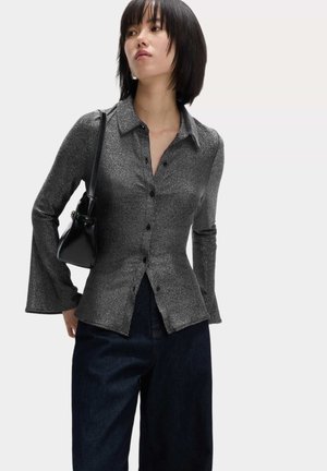 Schwarze, glänzende Hemdbluse mit langen, ausgestellten Ärmeln, kragenförmigem Design und dezentem Glitzer, kombiniert mit einer dunklen Jeans und einer kleinen schwarzen Tasche.