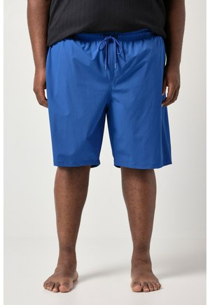 Mężczyzna w jasnoniebieskich spodenkach z troczkiem i czarnej koszulce, stojący boso na jednolitym jasnym tle.