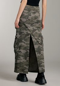 Camo maxi nederdel i grøn og grå, lavet af stof med en let glans. Har en frontslids og sidelommer. Bæres med sorte støvler.