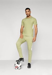 Muž v svetlozelenom športovom oblečení s čiernymi kopačkami stojí s jednou nohou na bielo-čiernej futbalovej lopte na jednoduchom pozadí.