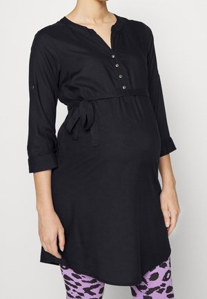 Schwarze Umstandstunika mit V-Ausschnitt, Knopfleiste, dreiviertel Ärmeln und Taillengürtel, kombiniert mit lila Leggings mit Leopardenmuster.