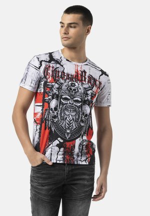 Jeune homme aux cheveux courts portant un t-shirt graphique blanc et rouge avec un motif de crâne viking et un jean foncé, posant avec une main dans la poche.