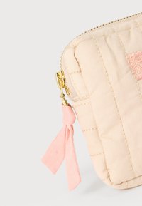 Μπεζ υφασμάτινο πουγκί με καπιτονέ σχέδιο, χρυστό φερμουάρ και ροζ υφασμάτινη ταινία στο φερμουάρ δεμένη σε κόμπο, σε λευκό φόντο.