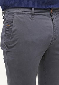Grijze katoenen broek met een slanke pasvorm, voorzien van een knoopsluiting, zijzakken en een subtiele textuur. Heeft een klein oranje detail bij de zak.