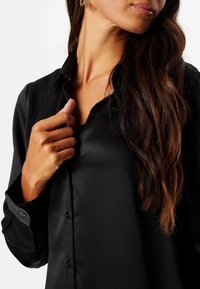 Blusa de satén negro con cuello, mangas largas y cierre de botones en la parte delantera. Presenta una textura suave y un ligero brillo.