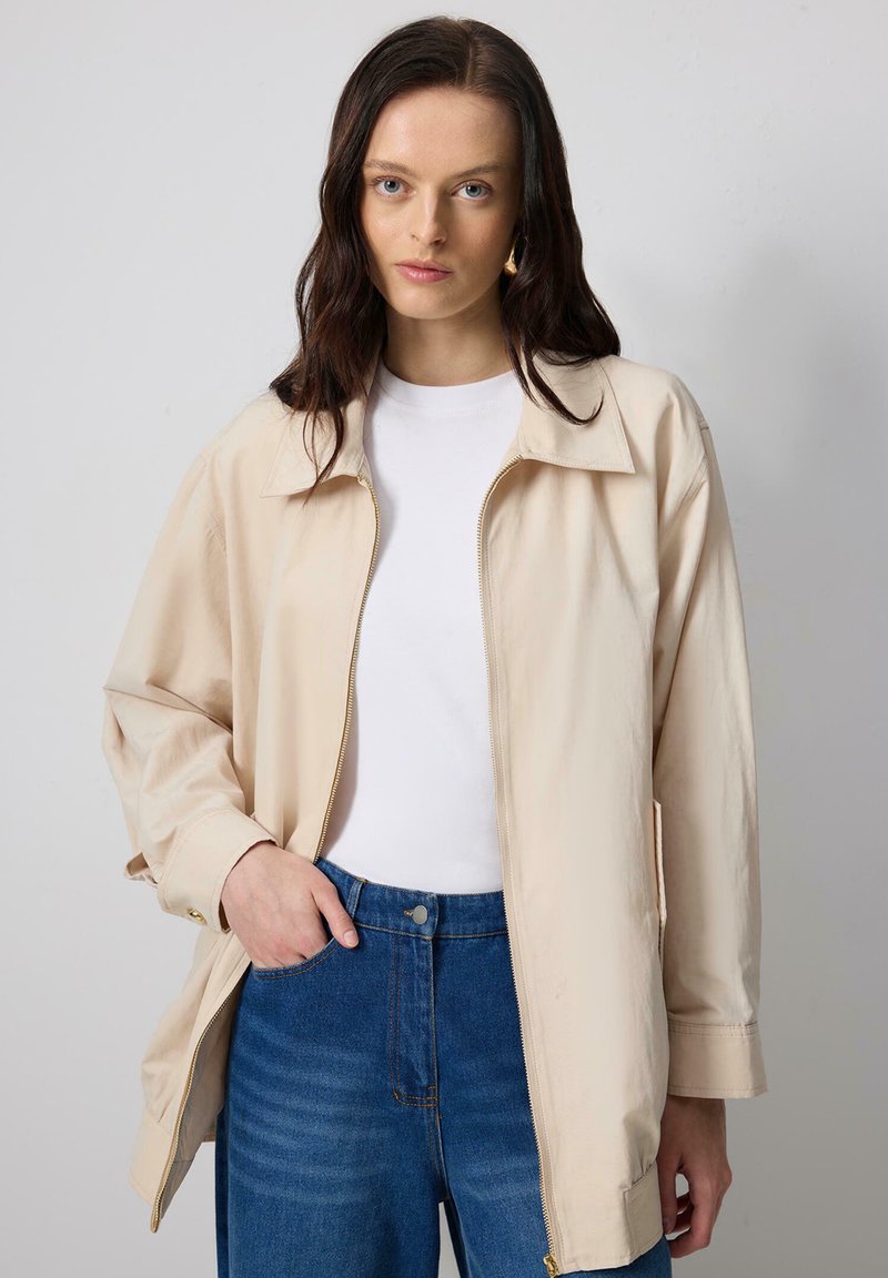 Beige zip-up jacket made of smooth, lightweight fabric; features a pointed collar and side pockets; paired with a white shirt and blue jeans.
