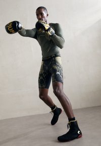 Boxer in una maglietta a maniche lunghe verde oliva aderente, pantaloni corti a compressione neri con motivo, e stivali neri con suole rosse, che indossa guanti neri.