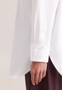 Camicia bianca a maniche lunghe con taglio dritto, dotata di polsini con tre bottoni e un tessuto liscio e leggero. Dettagli di cucitura visibili ai lati.