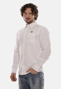 Camicia bianca a maniche lunghe con colletto button-down, dotata di taschino sul petto e piccolo logo ricamato sul lato sinistro. Texture liscia.