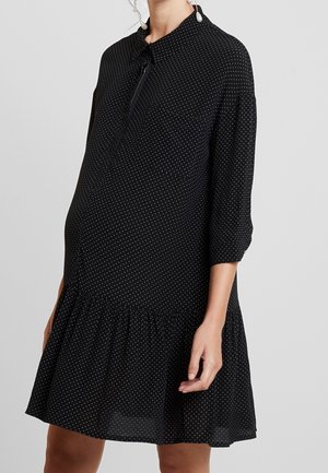 Robe de maternité noire à pois blancs, manches trois-quarts, ourlet volanté et col à revers sur une femme debout.