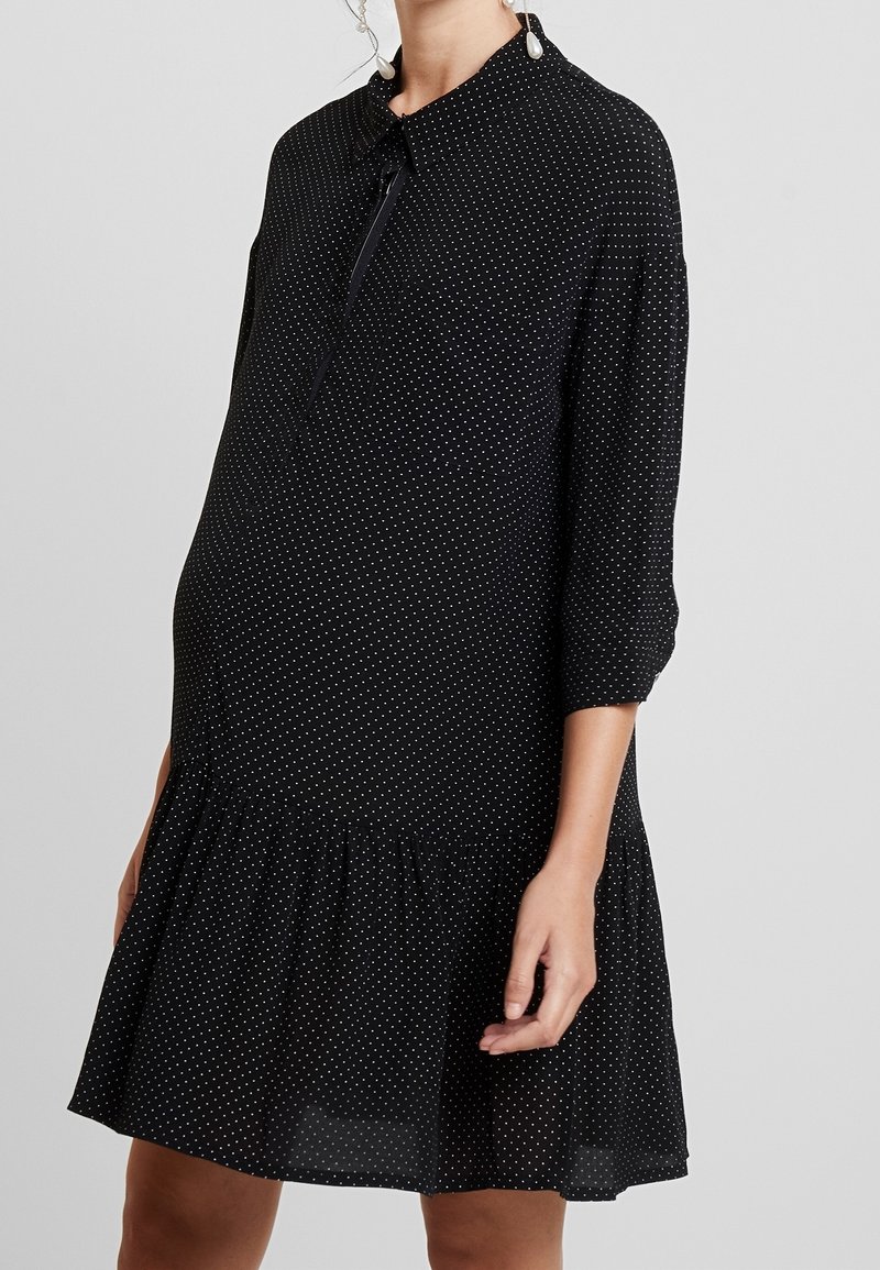 Robe de maternité noire à pois blancs, manches trois-quarts, ourlet volanté et col à revers sur une femme debout.