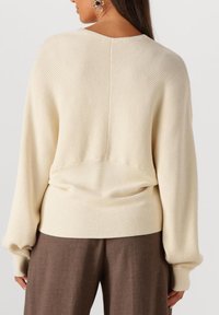 Frau in cremefarbigem, geripptem Strickpullover mit langen Puffärmeln und braunen, weit geschnittenen Hosen, von hinten gezeigt.