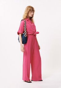 Roze, gestructureerde tweedelige outfit met een geknoopte short-sleeve blouse en wijde broek. Accessoires omvatten een blauwe geweven schoudertas.