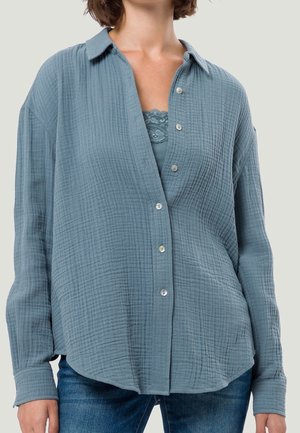 Mujer con una camisa azul suelta y texturizada de botones sobre un top azul con detalles de encaje, combinada con vaqueros azules sobre un fondo claro.