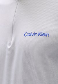 Ανοιχτό γκρι πόλο μπλούζα με υφή. Διαθέτει μισό φερμουάρ και μπλε λογότυπο "Calvin Klein" εμφανώς τοποθετημένο στο στήθος.