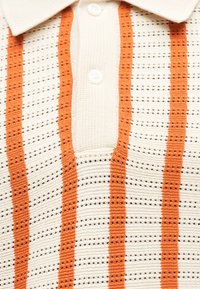 Gros plan d'un polo en maille blanc avec des rayures verticales orange et des boutons blancs sur la patte de boutonnage avant.