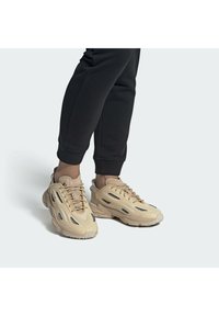 Osoba v béžových chunky teniskách s černými síťovanými detaily a černých joggingových kalhotách, stojící na jednoduché světle šedé podlaze.