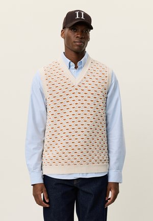 Pletená krémová vesta s oranžovým diamantovým vzorom, s výstrihom do V, nosená cez svetlomodrú košeľu; kombinovaná s tmavými džínami a hnedou čiapkou.