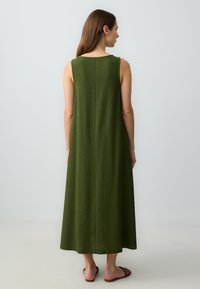 Kobieta z długimi brązowymi w�łosami, ubrana w bezrękawową oliwkowozieloną sukienkę midi i brązowe sandały na płaskiej podeszwie, stojąca na tle jasnego, jednolitego tła.