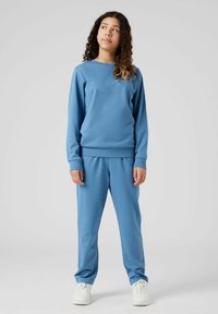 Gai zils krekls ar garām piedurknēm un atbilstošas bikses, kas izgatavotas no mīkstas auduma. Vienkāršs dizains ar ribotiem piedurkņu galiem un jostasvietu. Baltas sporta kurpes noslēdz apģērbu.