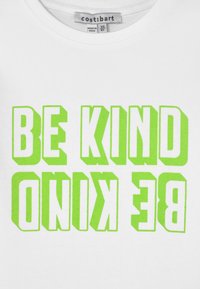 T-shirt de algodão branco com texto verde em negrito e invertido que diz "SEJA GENTIL" em uma fonte alta e em maiúsculas. A etiqueta mostra o nome da marca.