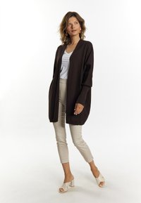 Bruine open voorkardigan van zachte brei, gecombineerd met lichtbeige aangesloten broek en crèmekleurige blokhak sandalen, tegen een neutrale achtergrond.