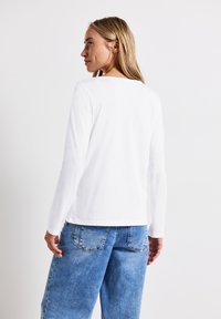 Hvid langærmet skjorte med rund hals, parret med lyseblå denimjeans. Stoffet ser blødt og let ud.