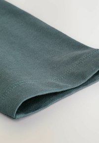 Prim-plan al unei mâneci pliate, realizată dintr-un material tricotat moale, de culoare teal, cu o cusătură dublă vizibilă, pe un fundal alb.