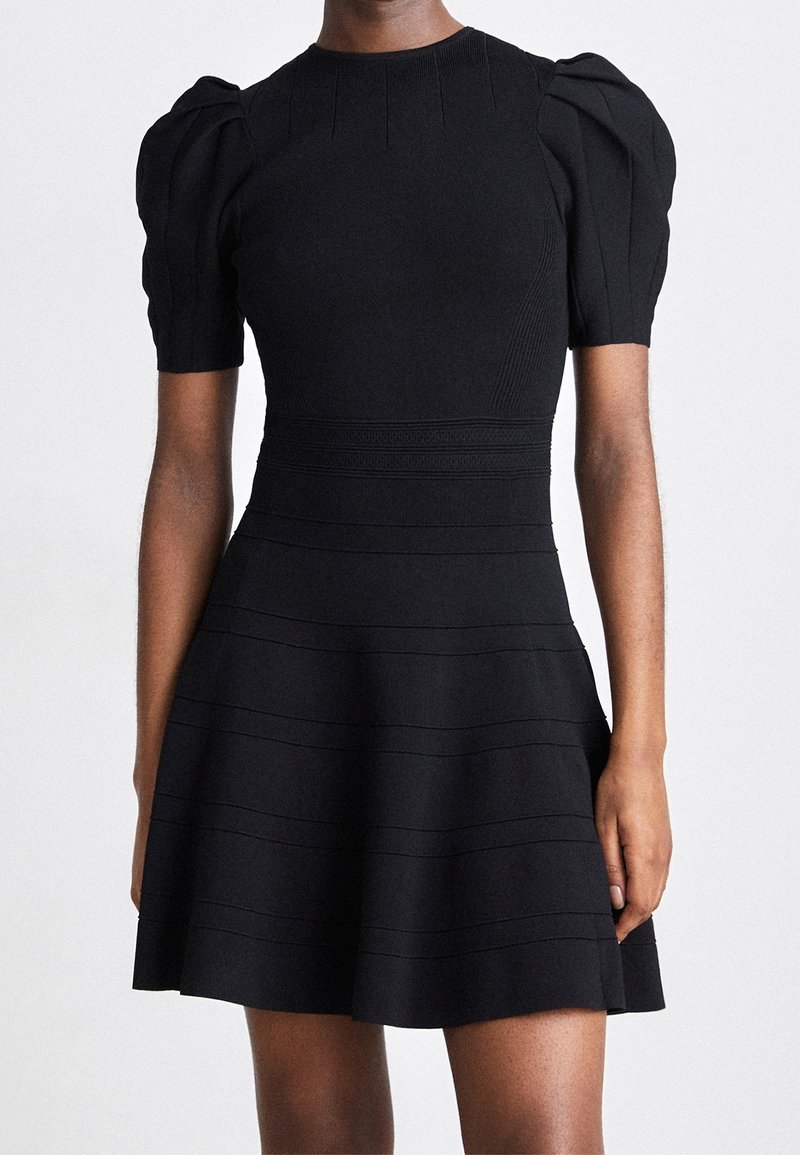 Robe noire à manches courtes avec épaules bouffantes, taille cintrée, tissu texturé et jupe évasée portée par une personne debout sur un fond uni.