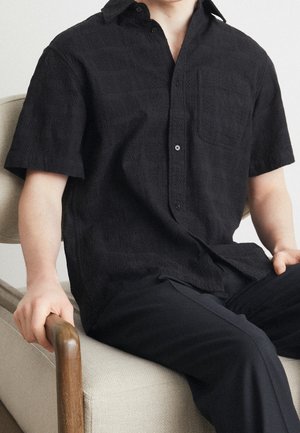 Schwarzes kurzärmliges Hemd mit einem strukturierten Muster, Knopfleiste und einer einzelnen Brusttasche, kombiniert mit dunklen Hosen. Auf einem hellen Sofa sitzend.
