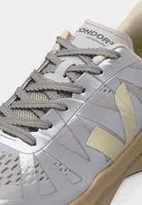 Κοντινό πλάνο γκρι και μπεζ αθλητικού παπουτσιού με πλέγμα στο επάνω μέρος, αντανακλαστικές επενδύσεις, ανάγλυφη σόλα και ετικέτα με την επιγραφή "CONDOR3 ADVANCED" στη γλώσσα.