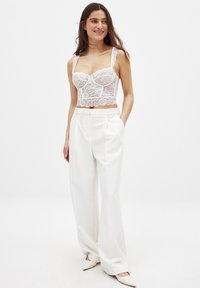 Top corto bianco in pizzo con spalline regolabili, caratterizzato da motivi floreali. Abbinato a pantaloni bianchi a vita alta e scarpe con tacco a punta.