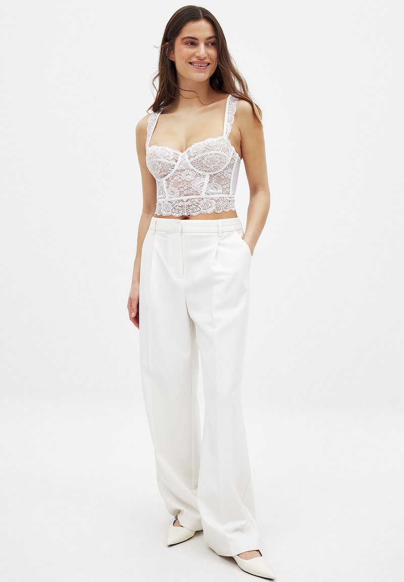 Top corto bianco in pizzo con spalline regolabili, caratterizzato da motivi floreali. Abbinato a pantaloni bianchi a vita alta e scarpe con tacco a punta.