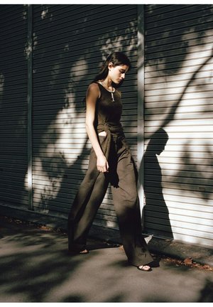 Young woman in dark sleeveless top and wide-leg pants walking by a metal shutter with tree shadows cast on it.