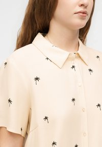 Γυναίκα που φοράει μπεζ πουκάμισο με κοντά μανίκια με μαύρα σχέδια φοίνικα και με μυτερό γιακά, με μακριά καστανά μαλλιά κάτω.